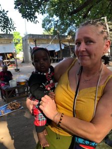 Une femme blonde, souriante, porte un petit bébé dans ses bras. Elle est habillée d’un débardeur jaune et a les cheveux tressés. Le bébé porte un vêtement sombre avec des touches rouges et regarde l’appareil photo. Ils se trouvent à l’extérieur, sous un grand arbre qui crée de l’ombre. En arrière-plan, on aperçoit des maisons simples et des femmes assises, probablement dans un cadre de vie villageois. L’ambiance est chaleureuse, paisible et humaine.