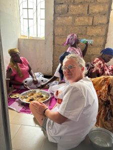 Plusieurs femmes sont assises au sol à l’intérieur d’une pièce simple aux murs clairs et partiellement en briques. Elles partagent un repas disposé dans un grand plat commun posé sur un tissu coloré. Au premier plan, une femme européenne en blouse blanche est assise parmi elles, souriante, tenant une cuillère. En arrière-plan, d’autres femmes mangent ou boivent de l’eau. La scène évoque un moment de convivialité, de respect mutuel et de vie partagée.