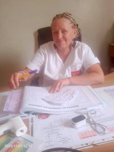 Une femme européenne portant une blouse blanche est assise à un bureau. Elle tient un stylo et remplit des documents médicaux étalés devant elle. On distingue des registres de consultation, des fiches de suivi, un tensiomètre et du matériel de soins. Elle regarde l’appareil photo avec un léger sourire. La scène se déroule dans un bureau simple, évoquant un centre de santé. L’image met en lumière le travail administratif et essentiel du suivi médical.