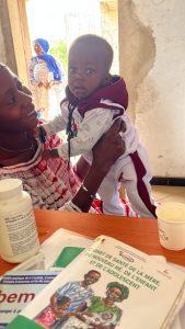 Une mère africaine tient son bébé debout sur une table, le regardant avec un sourire tendre. Le bébé, habillé d’une tenue claire avec une capuche foncée, regarde l’appareil photo avec curiosité. Sur la table, on voit des documents médicaux, notamment un carnet de santé de la mère et de l’enfant, ainsi que des médicaments et un gobelet. En arrière-plan, une autre femme se tient près d’une ouverture, laissant entrer la lumière extérieure. La scène se déroule dans un centre de santé, dans une atmosphère chaleureuse et rassurante.