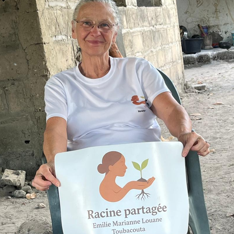 Marianne, co-fondatrice de Racine Partagée, assise en extérieur sur un sol sablonneux au Sénégal, tenant fièrement une affiche avec le logo de l'association et les prénoms des fondatrices.