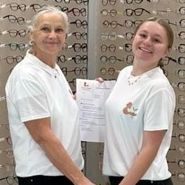 Marianne, trésorière, et Louane, secrétaire de l'association Racine Partagée, posent ensemble dans un magasin d'optique, tenant un document administratif et un sac, symbolisant la collaboration intergénérationnelle.