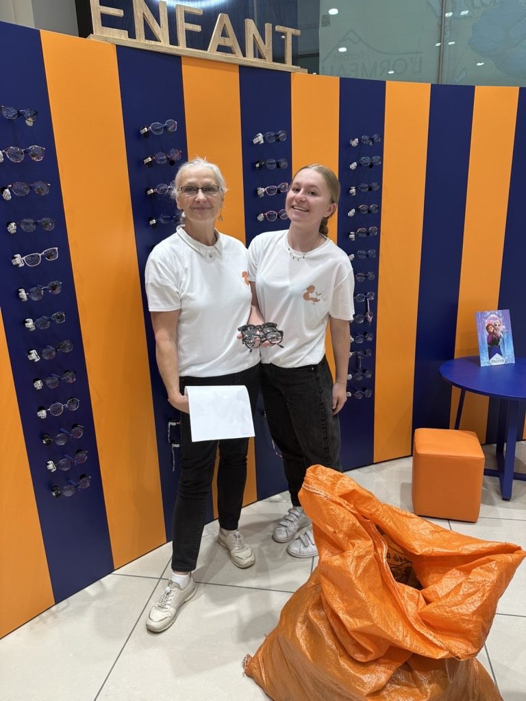 Deux femmes se tiennent côte à côte dans un espace intérieur. Elles portent des tee-shirts clairs et se trouvent devant un mur présentant de nombreuses paires de lunettes exposées. L’une d’elles tient plusieurs lunettes dans ses mains. Un grand sac est posé au sol, au premier plan. Le lieu est lumineux et organisé.
