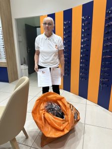 Parfait, même format 🌿 Titre Rassembler pour transmettre Description aveugle Une femme adulte se tient debout dans un espace intérieur. Elle tient des documents dans ses mains. À ses pieds, un grand sac ouvert contient plusieurs objets. Derrière elle, des murs présentent de nombreuses paires de lunettes exposées. Le lieu est lumineux et ordonné.