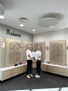 Deux femmes adultes se tiennent debout dans un magasin d’optique. Elles se font face et tiennent ensemble un document et un petit sac. Autour d’elles, des présentoirs muraux exposent de nombreuses paires de lunettes. L’espace est clair, ordonné et éclairé par une lumière douce.