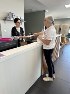 Deux femmes adultes se font face de part et d’autre d’un comptoir dans un magasin d’optique. L’une, derrière le comptoir, tient un document qu’elle remet à l’autre. Autour d’elles, le lieu est calme, lumineux, avec des étagères, un ordinateur, des présentoirs et un espace d’accueil ordonné.