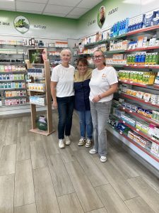 Trois femmes adultes se tiennent côte à côte dans un espace lumineux, entourées d’étagères remplies de produits de santé et de bien-être. Elles sourient face à l’objectif. Le lieu est organisé, chaleureux, avec une dominante de couleurs vertes et naturelles qui évoquent le soin, la prévention et l’accompagnement.