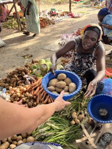 Une femme africaine est assise au sol dans un marché en plein air. Elle tend un grand bol bleu rempli de pommes de terre vers une main qui arrive au premier plan. Autour d’elle, des tas de légumes sont disposés : des patates douces, des racines et des feuilles vertes. Elle porte un foulard sur la tête et une robe à motifs. En arrière-plan, on distingue d’autres vendeurs et produits étalés au sol. L’ambiance reflète un moment de commerce simple, humain et chaleureux.