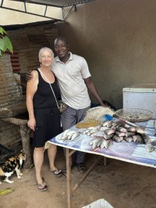 Une femme européenne aux cheveux courts et gris clair pose souriante aux côtés d’un homme africain. Il a son bras posé sur son épaule dans un geste amical. Ils se tiennent à l’intérieur d’un abri simple, probablement un lieu de travail artisanal. Devant eux, une table en bois est couverte de poissons fumés et séchés. À côté, on distingue un petit congélateur et des outils de préparation. L’ambiance est chaleureuse et authentique, évoquant un moment de partage dans la vie quotidienne du village.