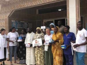 Un groupe de femmes et d’hommes est réuni devant l’entrée d’un centre de santé. Plusieurs personnes portent des blouses médicales, d’autres des vêtements traditionnels. Chacun tient du matériel médical ou des boîtes de soins. Au centre du groupe, une femme présente une pancarte avec le logo et le nom de l’association Racine Partagée. Tous regardent l’appareil photo avec des expressions sérieuses ou souriantes. La scène se déroule à l’extérieur du bâtiment, illustrant une action collective et solidaire au service de la santé.