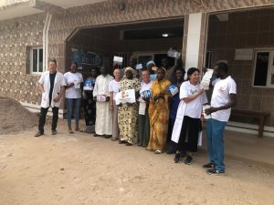 Un groupe de femmes et d’hommes est rassemblé devant un bâtiment de santé. Certaines personnes portent des blouses blanches, d’autres des vêtements traditionnels ou des t-shirts associatifs. Plusieurs tiennent dans leurs mains des boîtes de matériel médical et des documents. Au centre du groupe, une femme tient une pancarte avec le nom de l’association Racine Partagée. Tous sourient et regardent l’appareil photo. La scène se déroule à l’extérieur, devant l’entrée du centre, et reflète un moment de collaboration et de réussite collective.