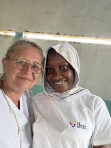 Deux femmes sourient côte à côte en regardant l’appareil photo. L’une est une femme européenne portant des lunettes et un haut blanc. L’autre est une femme africaine portant un voile clair brodé et un t-shirt blanc avec le logo du Fonds mondial. Elles sont à l’intérieur d’un bâtiment simple, aux murs clairs. Leurs visages sont proches, exprimant la complicité, la bienveillance et un engagement commun.