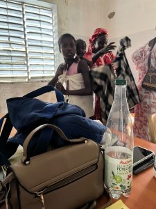 Au premier plan, une table sur laquelle sont posés un sac, un téléphone et une bouteille d’eau. En arrière-plan, à l’intérieur d’une pièce aux murs clairs, une fillette africaine se tient debout, un tissu clair noué autour de son torse. Un jeune enfant est porté sur son dos. Derrière elle, une femme ajuste des vêtements, tenant un tissu à carreaux. D’autres femmes se tiennent à proximité. La scène évoque un moment de préparation, probablement après une consultation ou un temps de soin.