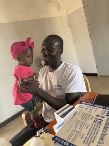 Un infirmier africain est assis derrière un bureau dans une salle de soins. Il porte un t-shirt blanc et tient doucement un bébé debout sur ses genoux. Le bébé est habillé en rose, avec un bandeau sur la tête, et regarde l’infirmier avec curiosité. Sur le bureau, on voit des boîtes de matériel médical, des seringues et des médicaments. La scène se déroule dans un espace simple, évoquant un centre de santé local. L’atmosphère est rassurante et bienveillante.
