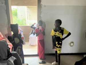 Plusieurs enfants africains se tiennent debout à l’intérieur d’un bâtiment clair. Certains tiennent des papiers ou des carnets dans leurs mains. Une fillette en tenue jaune et noire est appuyée contre un mur, une main sur la hanche. D’autres enfants sont regroupés près d’une porte ouverte qui donne sur l’extérieur. À l’arrière-plan, des adultes sont assis, attendant également. La scène évoque un moment d’attente dans un centre de santé, vu à hauteur d’enfants.
