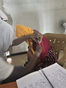Une soignante administre une injection dans le bras d’une femme assise sur une chaise en plastique. La patiente porte un vêtement rouge et jaune et détourne légèrement la tête pendant le soin. Sur la table à côté, un cahier ouvert avec des notes manuscrites et un stylo sont visibles, servant au suivi médical. La scène se déroule dans une pièce simple aux murs clairs, évoquant un centre de santé communautaire. L’image capture un moment précis de vaccination ou de soin préventif.