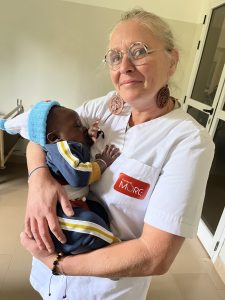 Une femme européenne portant une blouse blanche tient dans ses bras un bébé africain endormi. Le bébé porte un petit bonnet bleu et une tenue sombre avec des bandes claires. La femme sourit doucement vers l’appareil photo. Ils se trouvent dans une pièce claire, probablement un centre de santé, avec un lit médical visible à l’arrière-plan. L’ensemble de la scène évoque la tendresse, le soin et la sécurité.