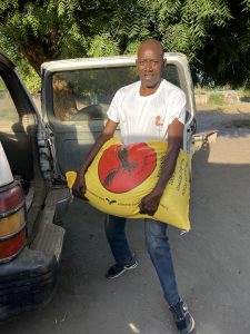 Un homme se tient à l’arrière d’un véhicule tout-terrain, dont la porte est ouverte. Il porte un t-shirt blanc avec le logo de l’association Racine partagée et un jean. Dans ses bras, il tient un grand sac jaune de riz, visiblement lourd. Il est debout sur un sol sablonneux, à l’ombre d’un arbre aux branches feuillues. La scène se déroule à l’extérieur, dans un environnement rural. L’image montre un moment de transport et de distribution, essentiel au soutien des familles.