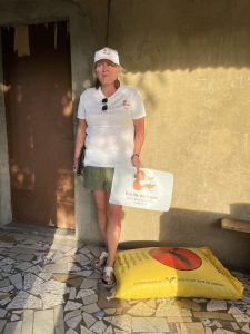 Une femme se tient debout contre un mur ocre, à l’extérieur d’un bâtiment simple. Elle porte un t-shirt blanc et une casquette avec le logo de l’association Racine partagée, ainsi qu’un short vert et des sandales. Dans sa main, elle tient une pancarte indiquant « Racine partagée – Emilie Marianne Louane – Toubacouta ». À ses pieds repose un grand sac jaune de riz. La lumière du soleil projette des ombres douces sur le mur, donnant à la scène une atmosphère calme et déterminée. L’image montre une présence engagée, ancrée dans le réel.