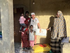 Deux femmes adultes, une adolescente et deux jeunes enfants sont rassemblés devant l’entrée d’une maison aux murs ocre, baignés par la lumière de fin de journée. L’une des femmes porte un jeune enfant dans ses bras. L’adolescente se tient près d’elle, droite et attentive. Un enfant plus jeune se tient au centre du groupe, face à l’objectif. À droite, une autre femme présente une pancarte portant le logo de l’association Racine partagée. Au sol, devant eux, repose un grand sac jaune de riz. La scène se déroule dans un cadre simple et intime, évoquant un moment de soutien et de présence familiale.