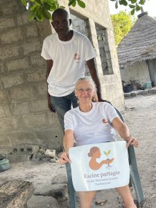 Deux adultes posent à l’extérieur, devant un bâtiment en pierre. Une femme est assise sur une chaise et tient une affiche sur laquelle est écrit « Racine partagée – Emilie Marianne Louane – Toubacouta », accompagnée d’un logo représentant une femme tenant une jeune plante. Un homme se tient debout derrière elle, posant une main sur le dossier de la chaise. Tous deux portent des t-shirts blancs avec le même logo. Autour d’eux, on distingue un environnement rural avec des arbres, une construction simple et un sol sablonneux. L’image évoque un partenariat humain et solidaire ancré dans un territoire.
