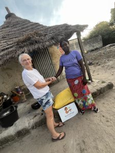 Deux femmes se tiennent face à face à l’extérieur, devant une habitation simple au toit de chaume. Elles se tiennent les mains, dans un geste à la fois symbolique et chaleureux. L’une porte un t-shirt blanc et un short, l’autre un t-shirt violet et une jupe longue colorée. À leurs pieds, un sac de denrées et une pancarte indiquant « Racine partagée – Emilie Marianne Louane – Toubacouta » sont posés sur le sol. Autour d’elles, on distingue des ustensiles de cuisine et un environnement rural, évoquant la vie quotidienne. La scène exprime un acte d’entraide directe et respectueuse.