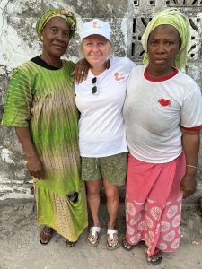 Trois femmes se tiennent debout, proches les unes des autres, devant un mur ancien. La femme au centre, à la peau claire, porte un t-shirt blanc et une casquette avec le logo de l’association Racine partagée. Elle entoure de ses bras deux femmes africaines, l’une vêtue de vert, l’autre de rose, toutes deux portant un foulard traditionnel. Les trois femmes regardent l’objectif avec des expressions calmes et dignes. Leur posture traduit une relation de respect, de soutien et de partenariat.