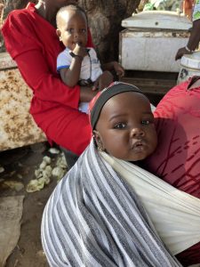 Un bébé africain est porté contre le dos d’une femme à l’aide d’un tissu rayé noir et blanc. Le bébé regarde vers l’appareil photo avec une expression calme. La femme porte un vêtement rouge. À l’arrière-plan, un autre jeune enfant est assis sur les genoux d’un adulte, également vêtu de rouge. La scène se déroule à l’extérieur, sous un arbre, avec un congélateur et des objets du quotidien autour. L’atmosphère évoque la vie de village, la famille et la protection des enfants.