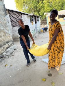 Deux jeunes femmes portent ensemble un grand sac jaune, visiblement lourd, tenu chacune par une extrémité. Elles sont debout dans une cour sableuse, devant des habitations modestes aux murs texturés. L’une porte une robe longue jaune et un foulard sombre, l’autre un t-shirt noir et un pantalon foncé. Elles sourient légèrement, concentrées sur leur effort commun. L’arrière-plan montre un environnement de vie quotidien, simple et vivant, avec des arbres et des objets domestiques.