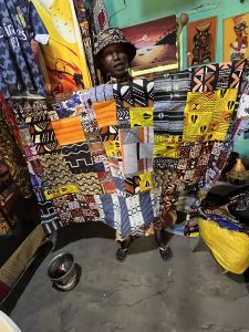 Une femme africaine se tient dans une petite boutique de tissus. Elle porte un chapeau à motifs et tient devant elle plusieurs pièces de tissu aux couleurs vives : jaune, bleu, noir et blanc, avec des motifs géométriques traditionnels. Autour d’elle, les murs sont décorés d’autres étoffes et de tableaux colorés. L’atmosphère évoque un lieu d’artisanat et de création, mettant en valeur le travail et la culture textile africaine.