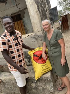 Un homme et une femme se tiennent côte à côte devant un mur ancien, aux pierres usées et fissurées. Entre eux, posé sur un muret, se trouve un grand sac jaune de riz, bien visible. L’homme, souriant, porte une chemise à carreaux et un short clair. La femme, calme et attentive, porte une robe verte et regarde l’objectif. La scène se déroule dans une ruelle ou une cour extérieure, dans un environnement simple et authentique, marqué par le quotidien.