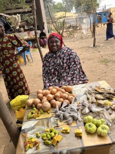Une femme africaine est assise derrière une petite table de vente en plein air. Elle porte un foulard rouge et un vêtement à motifs sombres. Elle sourit chaleureusement vers l’appareil photo. Sur la table devant elle, on voit des produits disposés à la vente : des poissons séchés, des légumes verts et des noix. En arrière-plan, le décor est celui d’un village : des arbres, des maisons colorées, quelques chaises en plastique et d’autres femmes qui vaquent à leurs occupations. L’atmosphère évoque la simplicité, le travail et la convivialité.