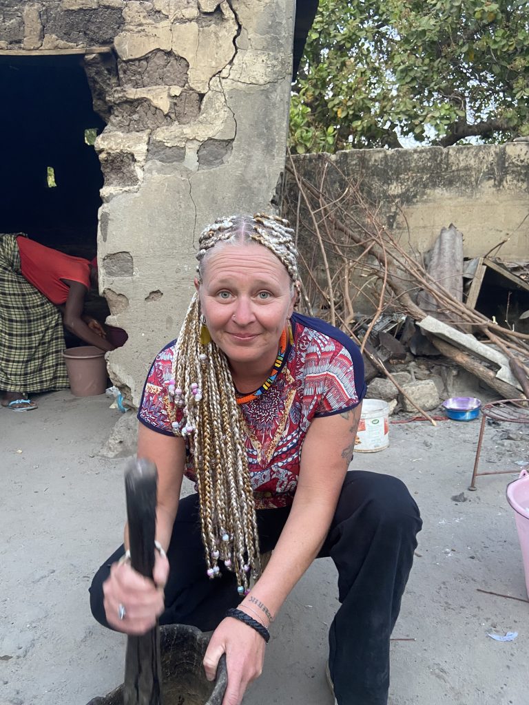 Émilie, présidente de Racine Partagée, coiffée de tresses, participe à la vie locale en pilant au mortier traditionnel dans un village au Sénégal.