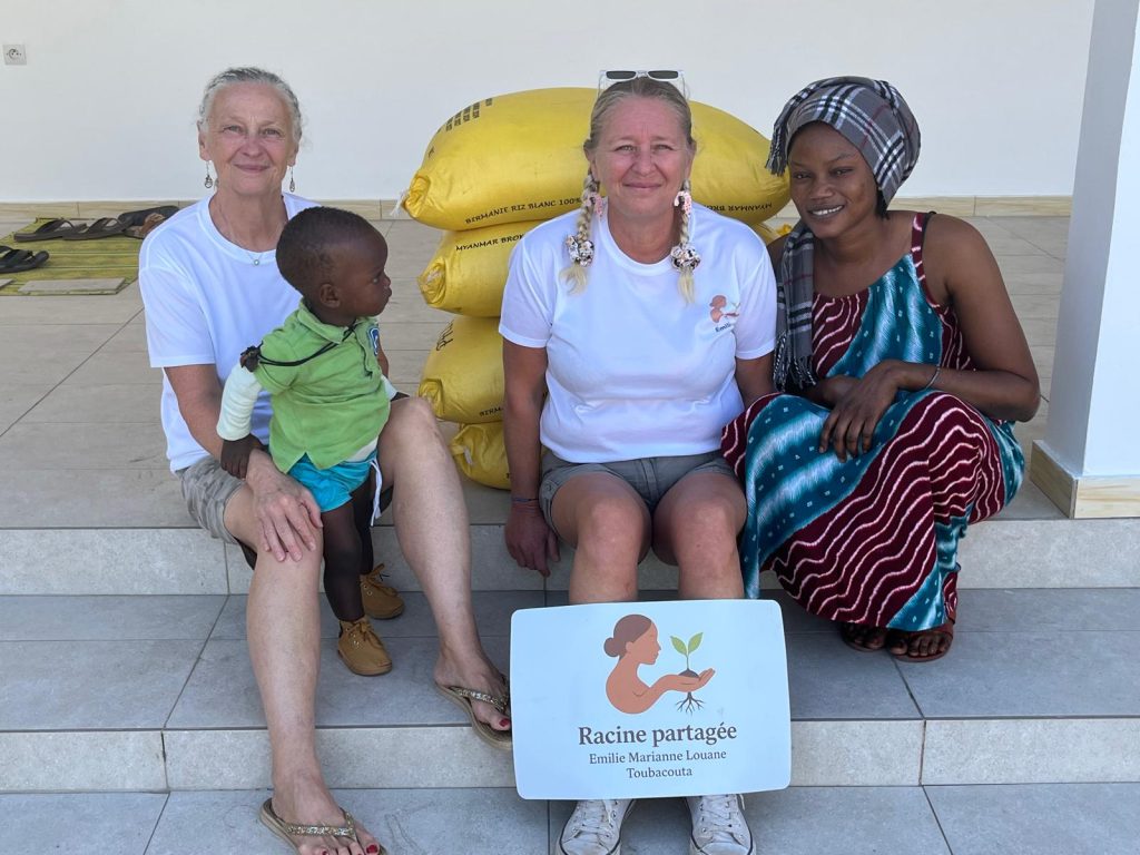 La présidente de l'association Racine Partagée et la trésorière posent assises sur des marches à Toubacouta, accompagnées d'une femme et d'un enfant du village. Elles sont entourées de sacs de vivres et tiennent le panneau du logo de l'association.